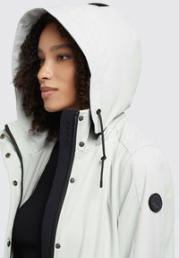 White waterproof jacket with a detachable hood, zip and snap closures, black accents, and textured interior lining. Features logo patch on shoulder.