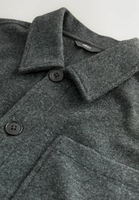 Sötétszürke gyapjú kabát strukturált gallérral, két gombbal és elülső zsebekkel. Az anyag puha, textúrált felületű, ami kiemeli a kivitelezést.