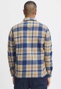 Gemberkleurig geruit flanellen shirt in beige, marineblauw en bruin; heeft een kraag, knoopmanchetten en een lus aan de achterkant. Zachte textuur.