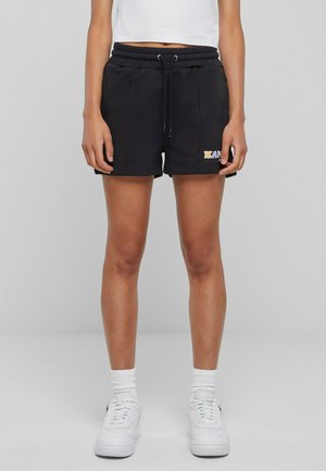 Vorderansicht einer Person, die schwarze Shorts mit Kordelzug und "KAN"-Logo, weißes kurzes Top, weiße Socken und weiße Turnschuhe vor einfachem Hintergrund trägt.
