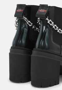 Sorte ankelstøvler med blank finish, chunky tekstureret sål, sølvkæde-detalje på anklen og logo-accenter ved kanten.