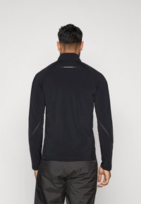 Cămașă sport neagră cu mâneci lungi și guler înalt, cu cusături plate și accente reflectorizante pe umeri și spate, fabricată dintr-un material elastic.