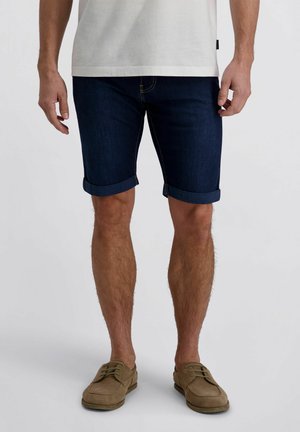 Mann trägt dunkelblaue, hochgekrempelte Jeansshorts, weißes T-Shirt und hellbraune Freizeitschuhe, steht vor einem einfarbigen weißen Hintergrund.