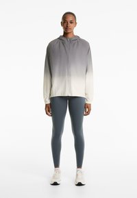 Giacca leggera con cappuccio in gradiente da grigio a bianco, con chiusura a zip e polsini elasticizzati, abbinata a leggings blu e sneakers bianche.