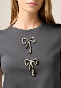 Top corto de manga corta en gris con dos apliques en forma de lazo joya, adornados con cristales claros y contornos negros, que proporcionan un contraste texturizado.