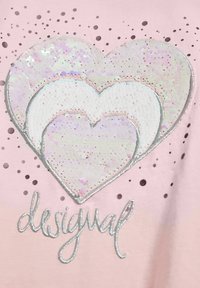 Tessuto rosa con un motivo di tre cuori ricoperte di paillettes di dimensioni diverse, bordate da cuciture argento, e la parola "desigual" in calligrafia argentata.