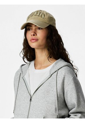 Frau mit welligem dunklem Haar, die eine beige Soho London Mütze, ein weißes Hemd und einen hellgrauen Reißverschluss-Hoodie trägt, leicht nach rechts blickend vor weißem Hintergrund.