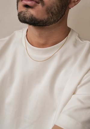 Weißes T-Shirt mit kurzen Ärmeln und weicher Textur, getragen mit einer dünnen goldenen Kettenhalskette. Nahaufnahme des Hals- und oberen Brustbereichs.