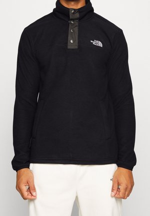 Mężczyzna noszący czarny polar z kołnierzem na guziki i białym logo The North Face na piersi, w połączeniu z białymi spodniami.