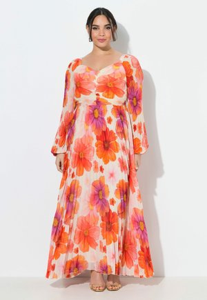 Kobieta w długiej, zwiewnej, białej sukience z dużym pomarańczowo-fioletowym kwiatowym wzorem, z długimi rękawami, w złotych sandałkach na otwartym czubku, stojąca na tle białej ściany.