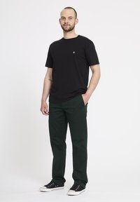 Zwarte katoenen t-shirt met ronde hals, gecombineerd met donkergroene broek en zwarte sneakers, tegen een eenvoudige witte achtergrond.