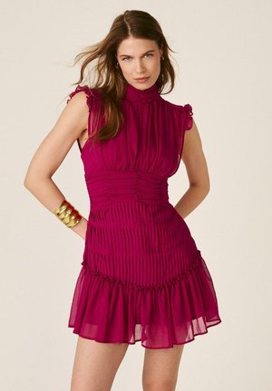 Mujer con vestido plisado sin mangas de color rosa oscuro con cuello alto y bajo con volantes, accesorio con pulsera de puño dorada, parada con las manos en las caderas.