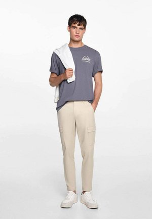 T-shirt en coton gris avec impression "VIVE SIMPLEMENT", pantalon cargo beige, baskets blanches, et un sweat-shirt blanc drapé sur l'épaule.