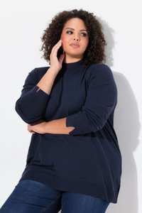Kvinde med krøllet hår iført navyblå sweater og jeans poserer med den ene hånd, der rører ved hendes ansigt, og den anden arm krydset over kroppen.