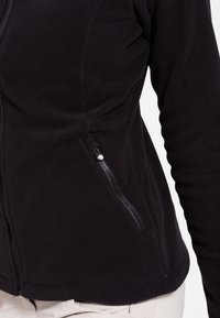 Musta fleece-takki, jossa on vetoketjullinen sivutasku, muotoon ommeltu malli, sileä rakenne ja pitkät hihat.