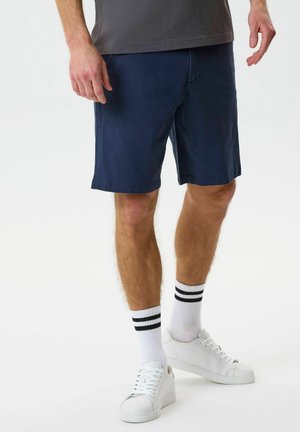 Férfi sötétkék rövidnadrágban, fekete csíkos fehér zokniban és fehér tornacipőben, egyszínű világos háttér előtt.