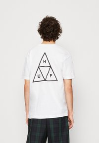 Camiseta de algodón blanca con un gran gráfico triangular negro en la parte posterior, con letras estilizadas 'H', 'U' y 'F'. Mangas cortas, ajuste relajado.