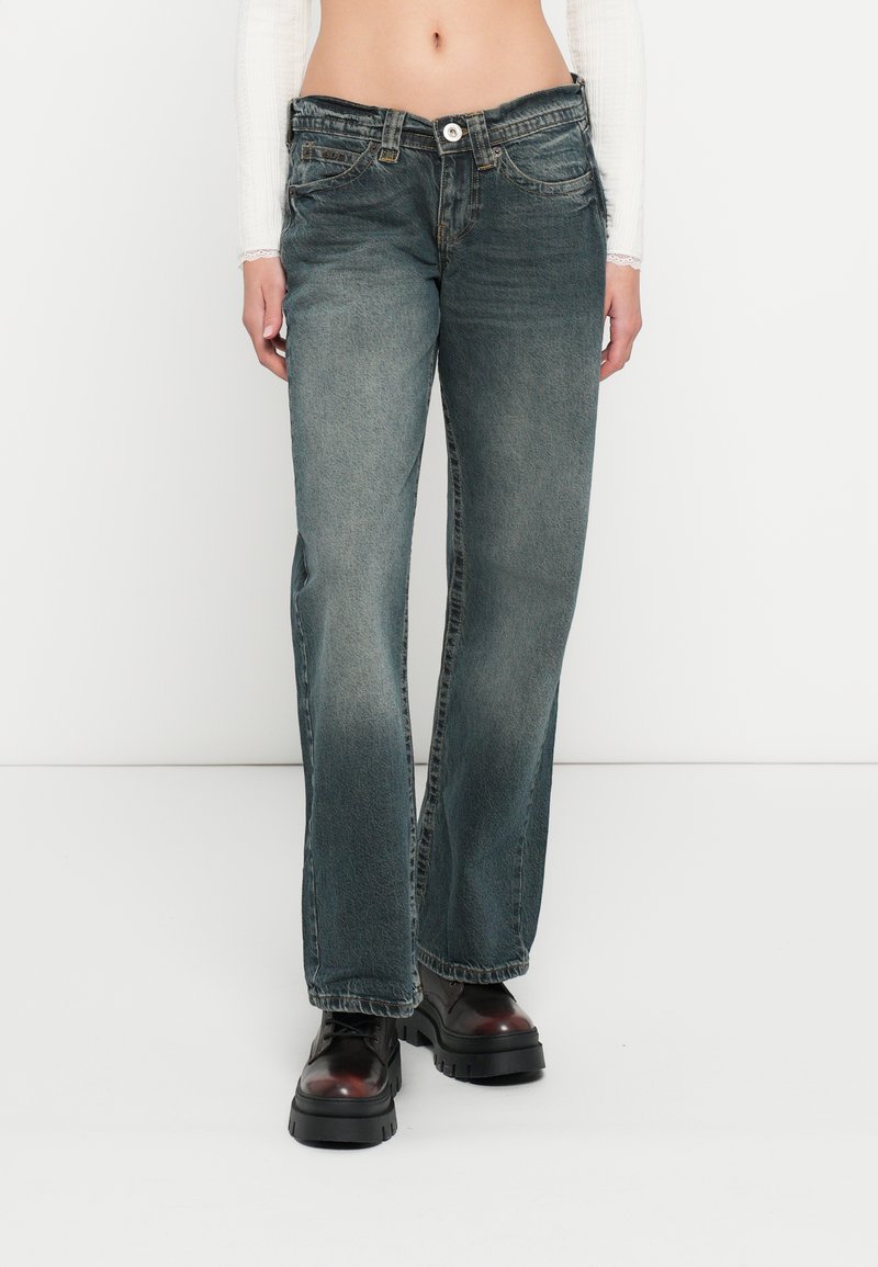 Jeans flare em azul desbotado feitos de ganga, com design de cinco bolsos e fecho de botão. Usados com botas pretas robustas.