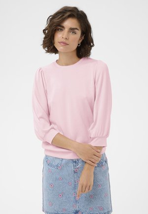 Femme aux cheveux courts et bouclés portant un haut rose pâle à manches bouffantes et une jupe en jean avec broderies florales roses, debout devant un fond blanc.