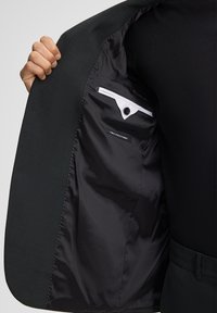 Zwarte blazer met een gestructureerde buitenzijde en een soepele, donkere voering. Het interieur heeft een knoopzakje en een label met de tekst "stretch stof."