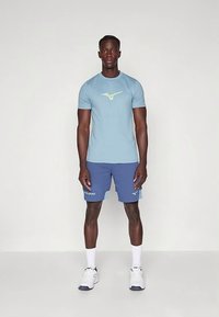 Šviesiai mėlyna sportinė marškinėliai su žaliu logotipu, derinami su mėlynais šortais, kuriuose yra šviesiai mėlyni akcentai, ir baltais sportiniais batais.