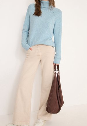 Femme portant un pull à col roulé bleu clair et un pantalon beige à jambes larges, tenant un sac marron avec des anses en anneau métallique dans la main droite.