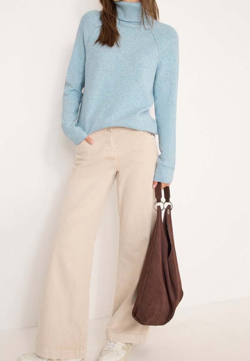Femme portant un pull à col roulé bleu clair et un pantalon beige à jambes larges, tenant un sac marron avec des anses en anneau métallique dans la main droite.