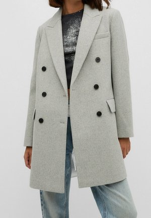 Femme portant un manteau en laine gris clair croisé sur un haut noir brillant et un jean bleu clair, debout devant un fond uni.