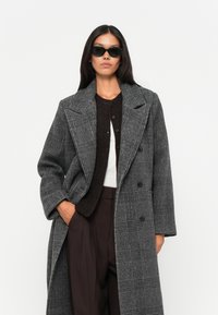 Grå, dobbeltradet frakke med ternet mønster, brede reverser og lange ærmer, parret med en mørk sweater og sorte solbriller.