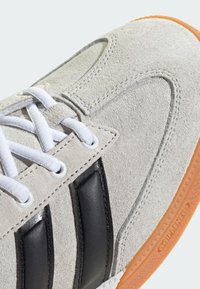 Semišové tenisky svetlo šedej farby s černými koženými pruhmi, bielymi šnúrkami a oranžovou gumovou podrážkou so štruktúrovanými vzormi a detailmi značky.