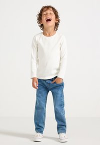 Witte ribbeltrui met lange mouwen gecombineerd met blauwe denim jeans. Het kind staat met de handen in de zakken, met witte sneakers op een effen achtergrond.