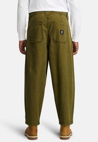 Olive green baggy pants with double back pockets and a logo patch, featuring a straight cut and cuffed hems, paired with tan footwear.