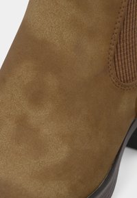 Gros plan d'une botte Chelsea en daim beige montrant le panneau latéral élastique, les détails de la couture et une partie de la semelle noire.