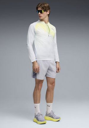 Ung mand iført hvid og gul lynlåstrøje, grå shorts, hvide Puma-sokker, grå og gule løbesko og store sorte solbriller, stående.