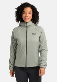 Giacca a zip verde chiaro con cappuccio. Presenta un logo sul lato sinistro del petto, una texture morbida e polsini elastici. Indossata sopra pantaloni scuri.