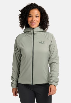 Veste zippée vert clair avec une capuche. Présente un logo sur la poitrine gauche, une texture douce et des poignets élastiques. Portée sur un pantalon foncé.