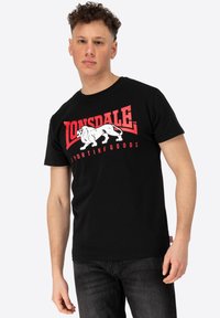 Juodas trumpomis rankovėmis marškinėliai su raudonu ir baltu "Lonsdale Sporting Goods" logotipu bei liūto grafika, atspausdinta priekyje. Minkštas medvilnės audinys.