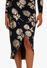 Donna che indossa una gonna nera con un motivo floreale metallico dorato e uno spacco frontale che mostra le sue gambe, con le mani appoggiate ai fianchi.