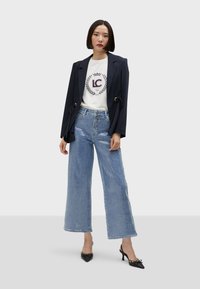 Navy blazer with gold accents, white t-shirt featuring logo, and light blue wide-leg jeans with a textured finish, paired with black heeled shoes.