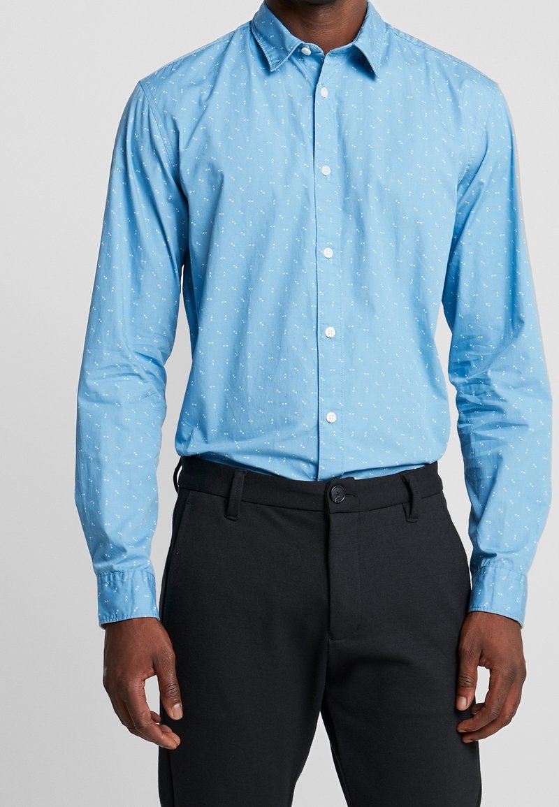 Muž oblečený v světle modré košili s dlouhým rukávem, se zapínáním na knoflíky a malými bílými vzory, a černých přizpůsobených kalhotách na jednoduchém pozadí.