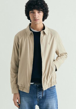 Joven con cabello rizado que lleva una chaqueta beige con cremallera, suéter negro, camiseta blanca interior y jeans azules, de pie contra un fondo claro sencillo.