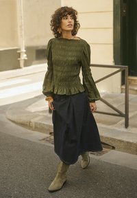 Frau mit lockigem Haar, die eine olivgrüne Rüschenshirt, einen dunklen Midi-Rock und passende grüne Stiefel trägt, während sie eine Stadtstraße entlanggeht.