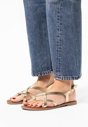 Gold strappy sandals feature a flat sole, crisscrossed metallic straps, and a buckle closure. Worn with cuffed blue jeans.