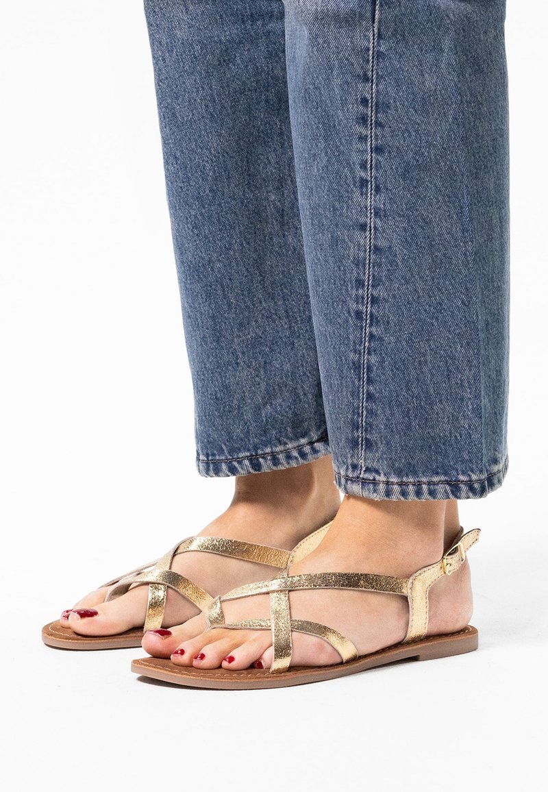 Gouden strappy sandalen hebben een platte zool, gekruiste metallic banden en een gespsluiting. Draag ze met opgerolde blauwe jeans.