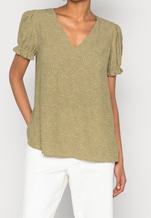 Blouse vert olive à manches courtes avec des pois blancs, col en V et poignets à volants portée avec un pantalon blanc.