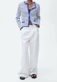 Cardigan a righe blu e bianco, con chiusura frontale a bottoni e due tasche, abbinato a pantaloni a gamba larga bianchi a vita alta e sandali neri.