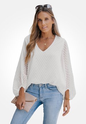 Witte top met chevronpatroon, lange doorschijnende mouwen en V-hals. Gecombineerd met lichtblauwe jeans en een beige clutch. Eenvoudige sieraden als accenten.