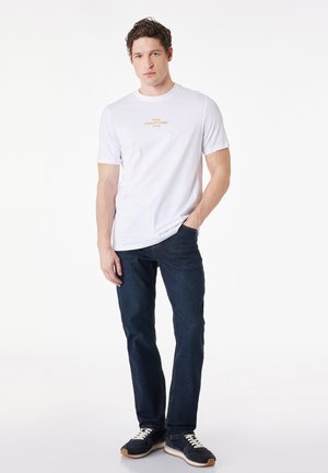 Mand stående med den ene hånd i lommen på mørke jeans, iført hvid t-shirt med orange tekst og sorte sneakers på hvid baggrund.