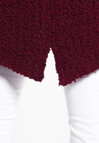 Bordeaux geklede trui met een unieke splitzoomdetail, weergegeven tegen witte broek. Het materiaal lijkt zacht en pluche.