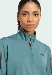 Teal athletic quarter-zip top made of smooth fabric. Features a high collar, black adidas logo on the chest, and black accents on the sleeves.